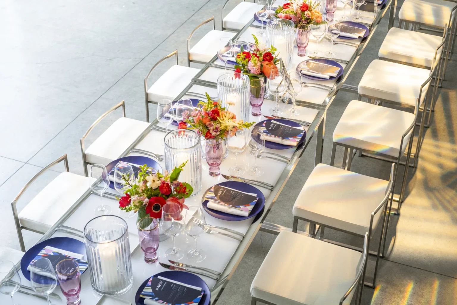 Elegant dining setup featuring a long glass table adorned with colorful floral arrangements and polished tableware.