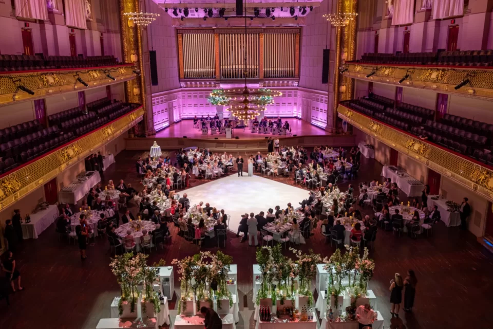 A grand event in a luxurious hall, featuring elegantly dressed guests dining and dancing under soft pink lighting and lush floral arrangements.
