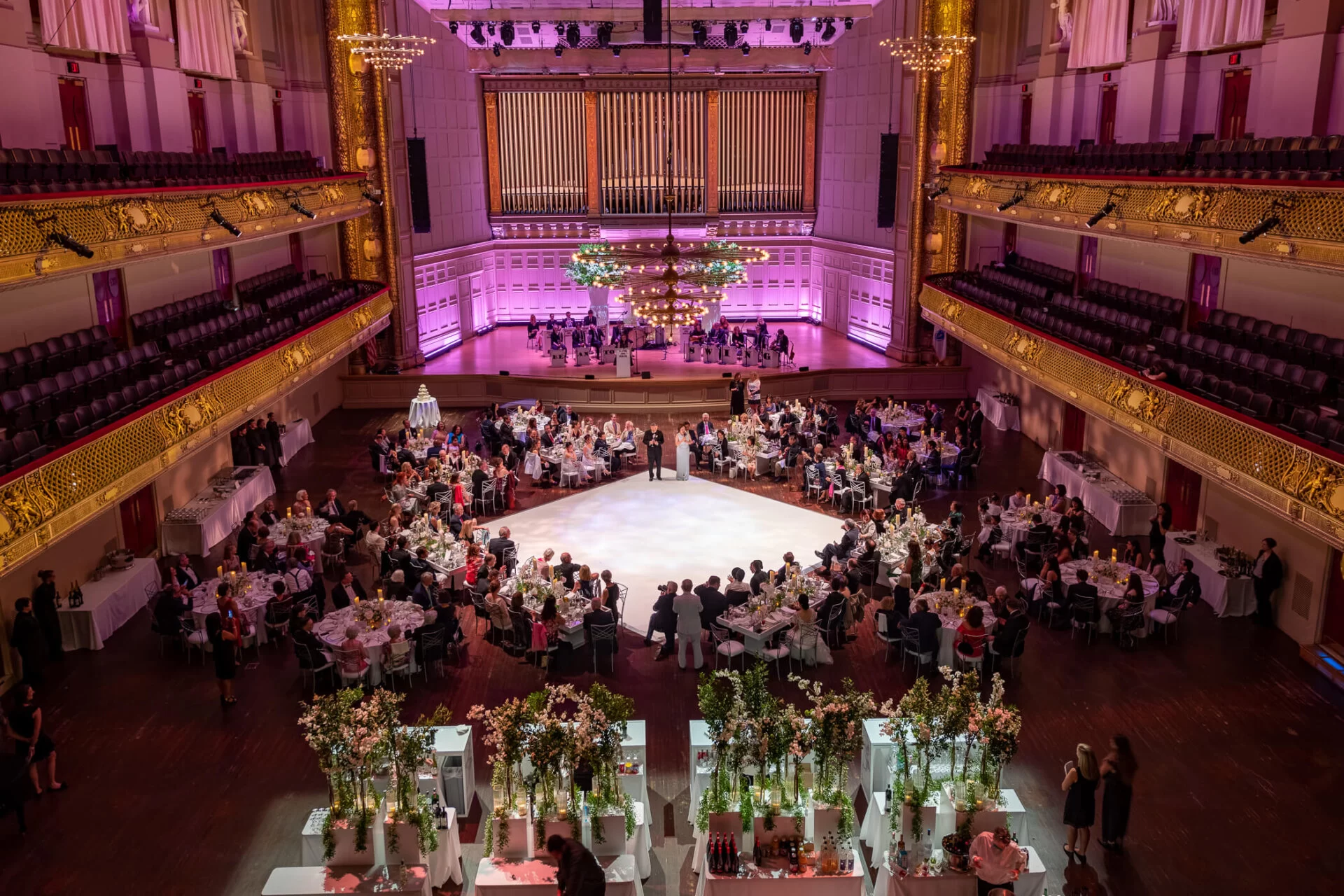 A grand event in a luxurious hall, featuring elegantly dressed guests dining and dancing under soft pink lighting and lush floral arrangements.