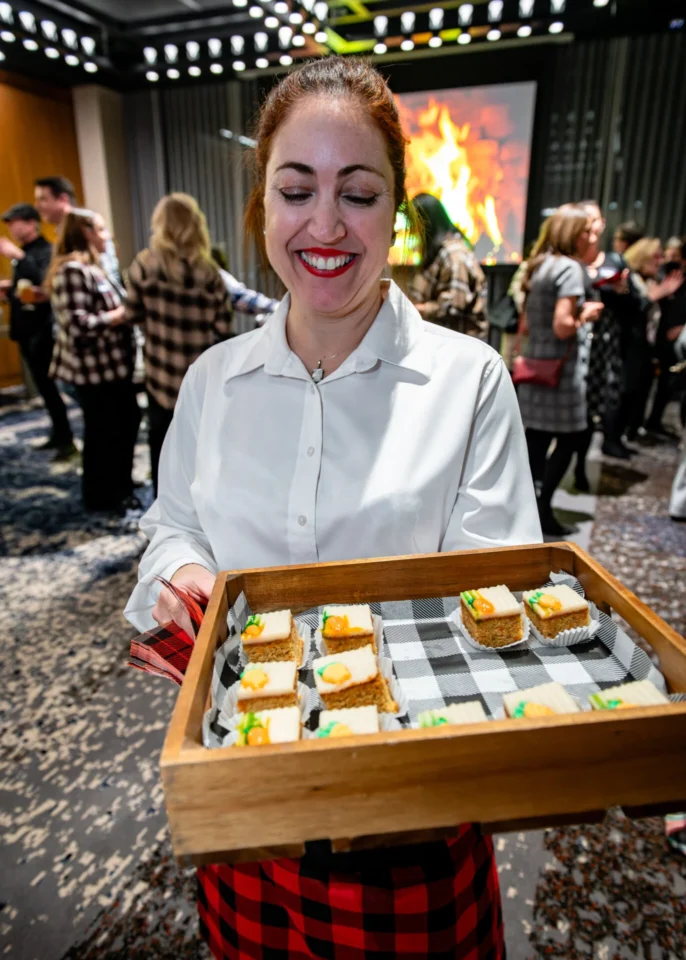 A person in a white shirt holds a wooden tray with decorated dessert squares, surrounded by a festive crowd in a stylish venue.