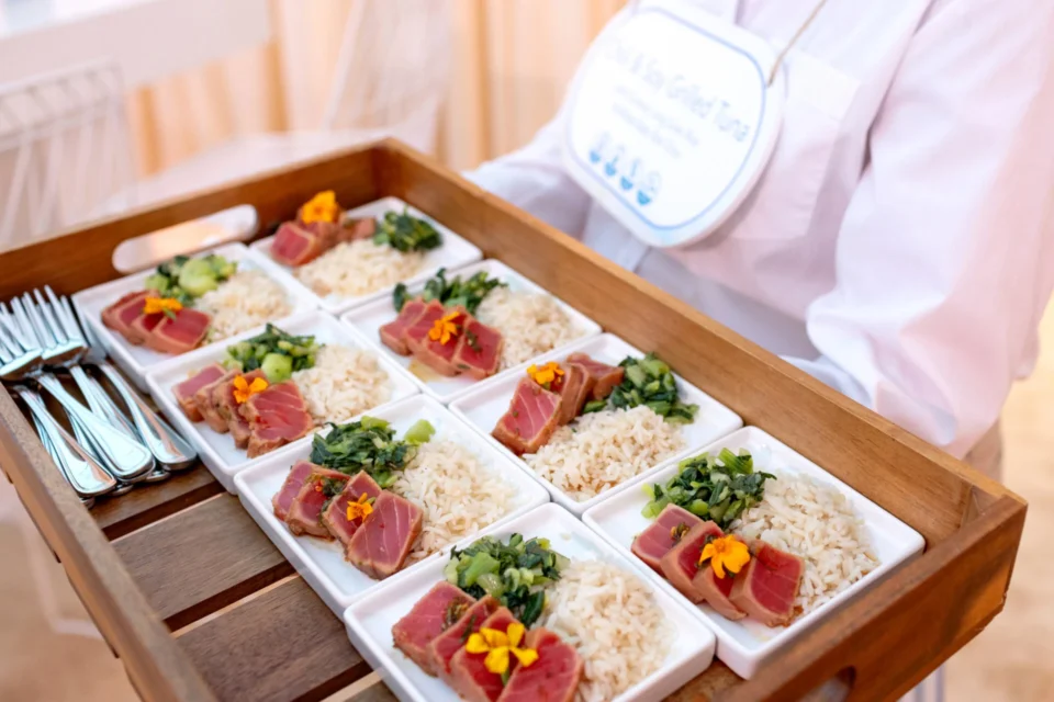 A server holds a wooden tray with neatly arranged plates of grilled tuna, rice, and greens, garnished with edible flowers.