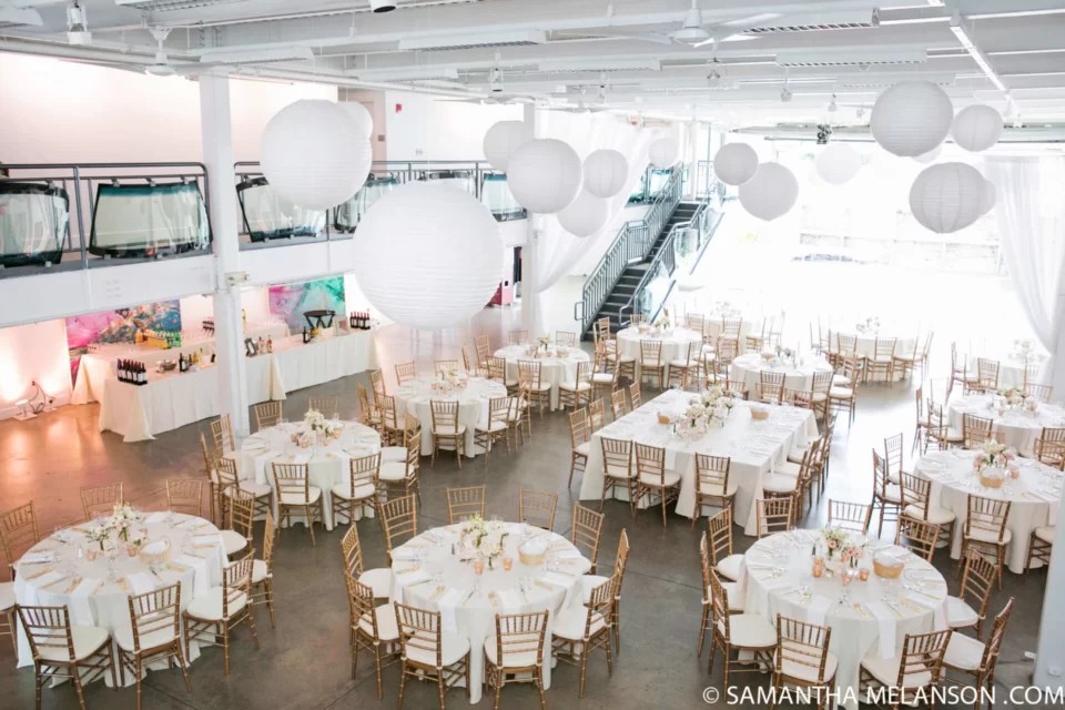 Elegant event space featuring round tables adorned with flowers, golden chairs, and hanging paper lanterns in a bright, airy atmosphere.