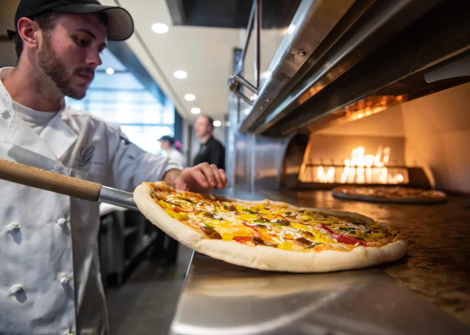 A chef carefully pulls a vibrant pizza topped with colorful vegetables from a wood-fired oven in a modern restaurant kitchen.