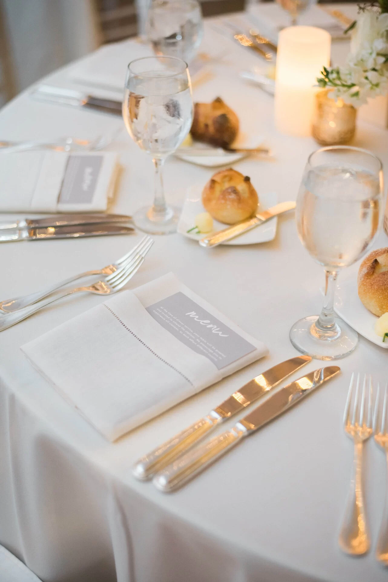 Elegant dining table setting with water glasses, silverware, a menu card, and freshly baked rolls on a white tablecloth.