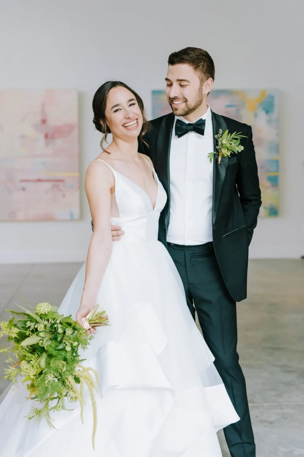 A bride in a flowing white gown holds a lush green bouquet, standing beside a groom in a classic tuxedo. Modern art is visible in the background.