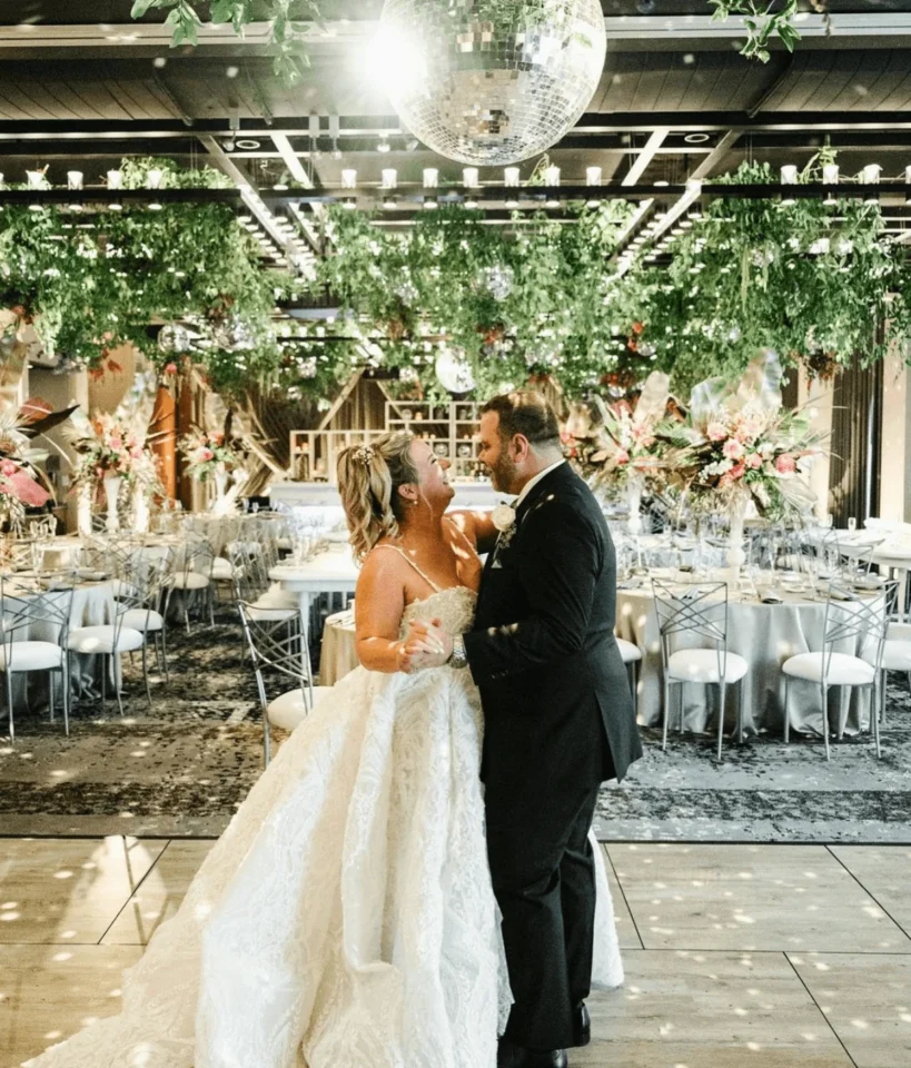A bride in an elegant lace gown and groom in a tuxedo embrace joyfully under a sparkling disco ball in a beautifully decorated reception hall.