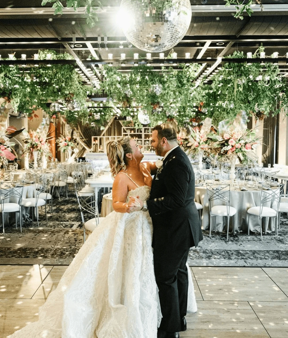 A bride in an elegant lace gown and groom in a tuxedo embrace joyfully under a sparkling disco ball in a beautifully decorated reception hall.